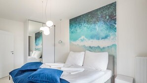 Et moderne soveværelse med en stor seng, hvidt sengetøj og en blå dekorativ pude. Der er et stort vindue med udsigt til himlen og et maleri på væggen.