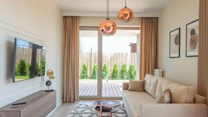 En moderne stue med sofa, sofabord og fjernsyn. Der er gardiner, et vindue med udsigt over grønne områder og en loftlampe.