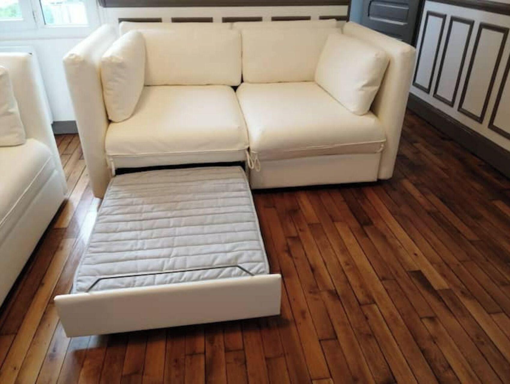 Een witte bank met uitschuifgedeelte, een houten vloer en een raam op de achtergrond.