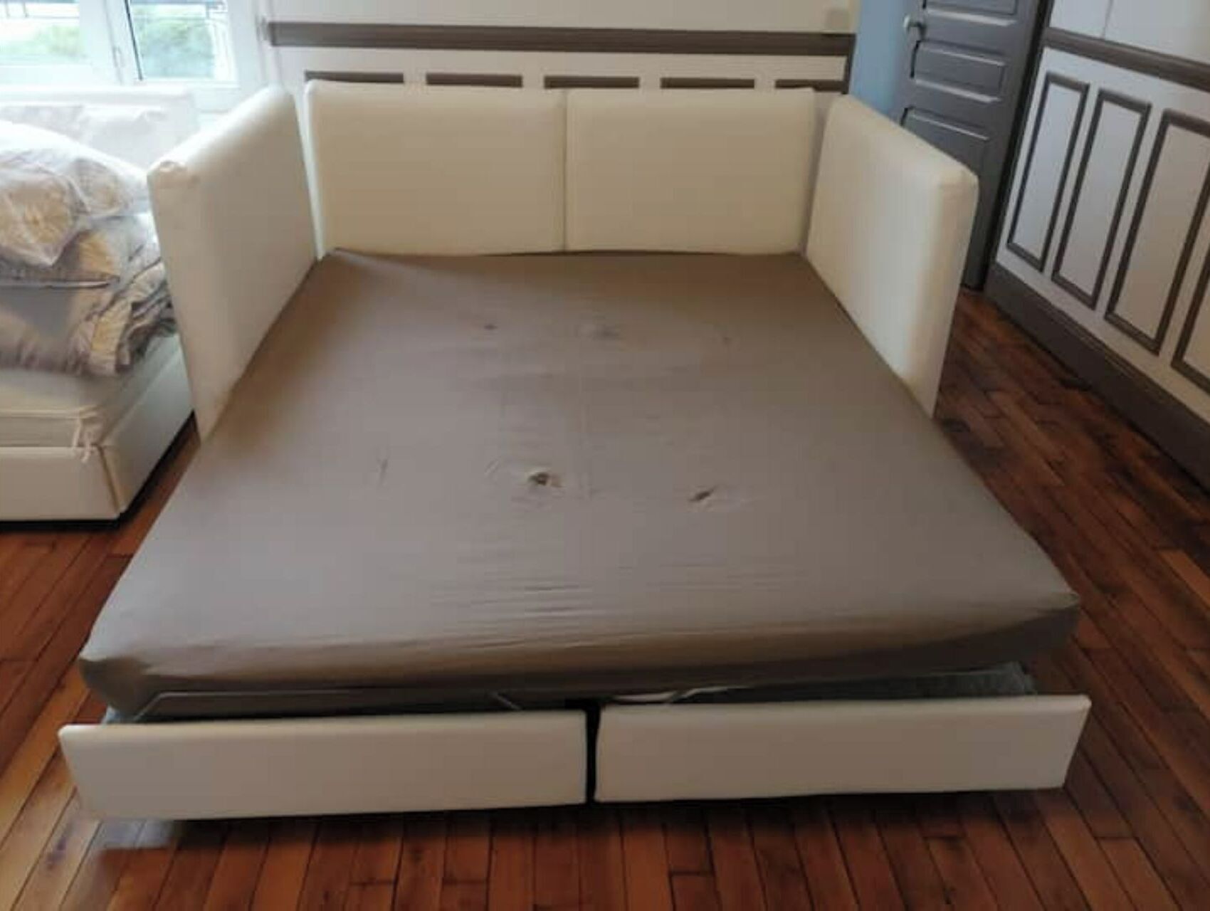 Una cama compacta con colchón y dos cajones laterales, suelo de madera y ventana con persianas.