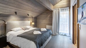 Quarto com teto de madeira, cama com roupa de cama branca e cinza, mesa de cabeceira, luminária fixada na parede e janela com cortinas.