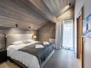 Ein Schlafzimmer mit Holzboden, einem Bett mit weißer und grauer Bettwäsche, einem Nachttisch, einer Wandlampe und einem Fenster mit Vorhängen.