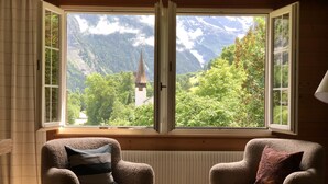 Une pièce avec deux fauteuils placés face à une grande fenêtre dont le volet est ouvert, offrant une vue sur une église et des montagnes.