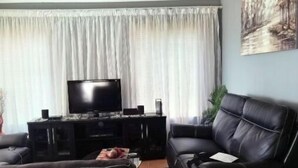 A living room with a black leather sofa, a flat-screen TV, and a large window with white curtains. - A home away from home (Mbombela)