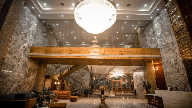 A hotel lobby with a grand chandelier, marble walls, a wooden reception desk, and a staircase. - Hotel Leroy Grand (Bengaluru)