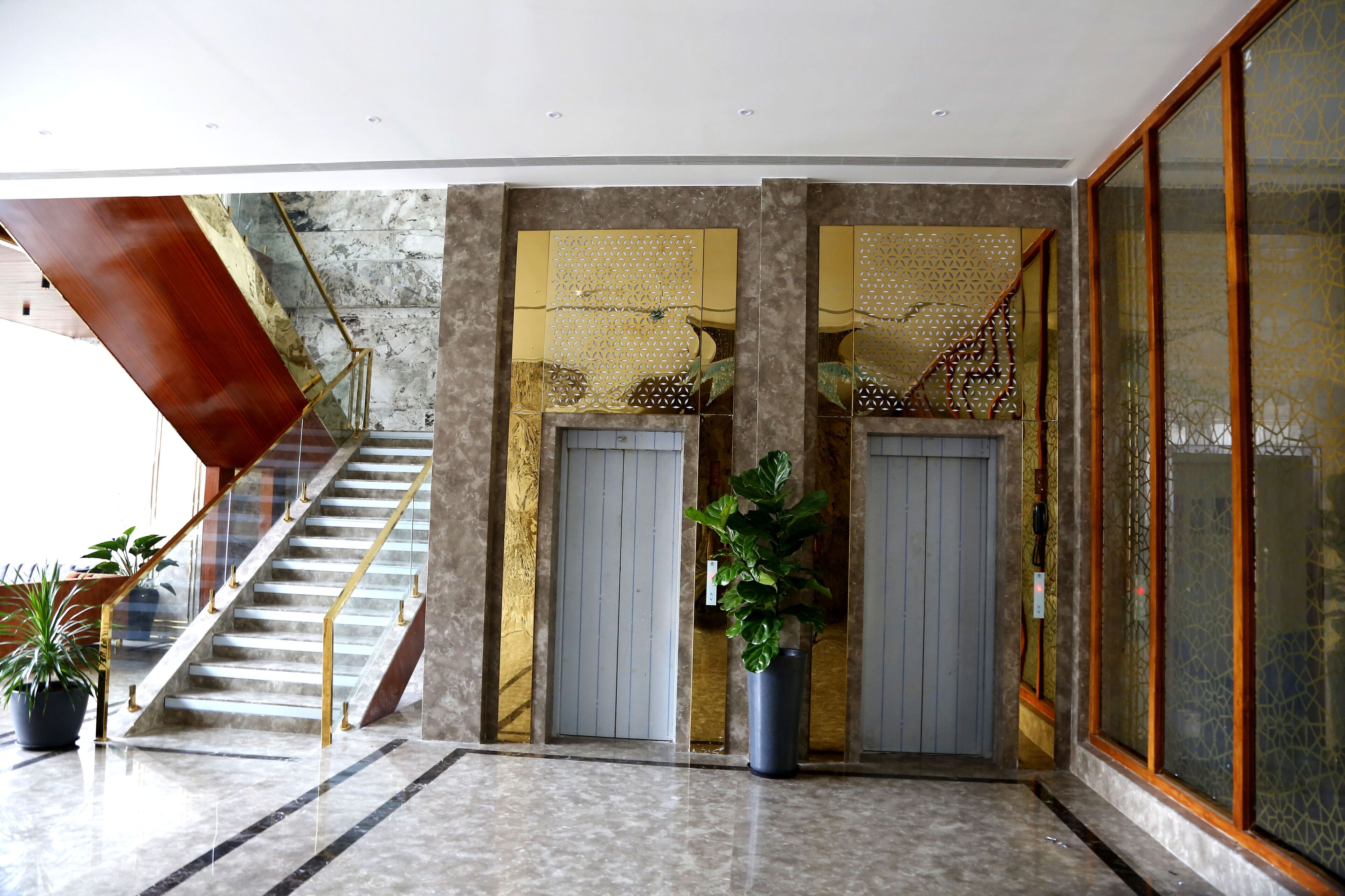 Rebedor modern d'hotel amb sòl de marbre, escala amb baranes daurades i una gran planta en test.