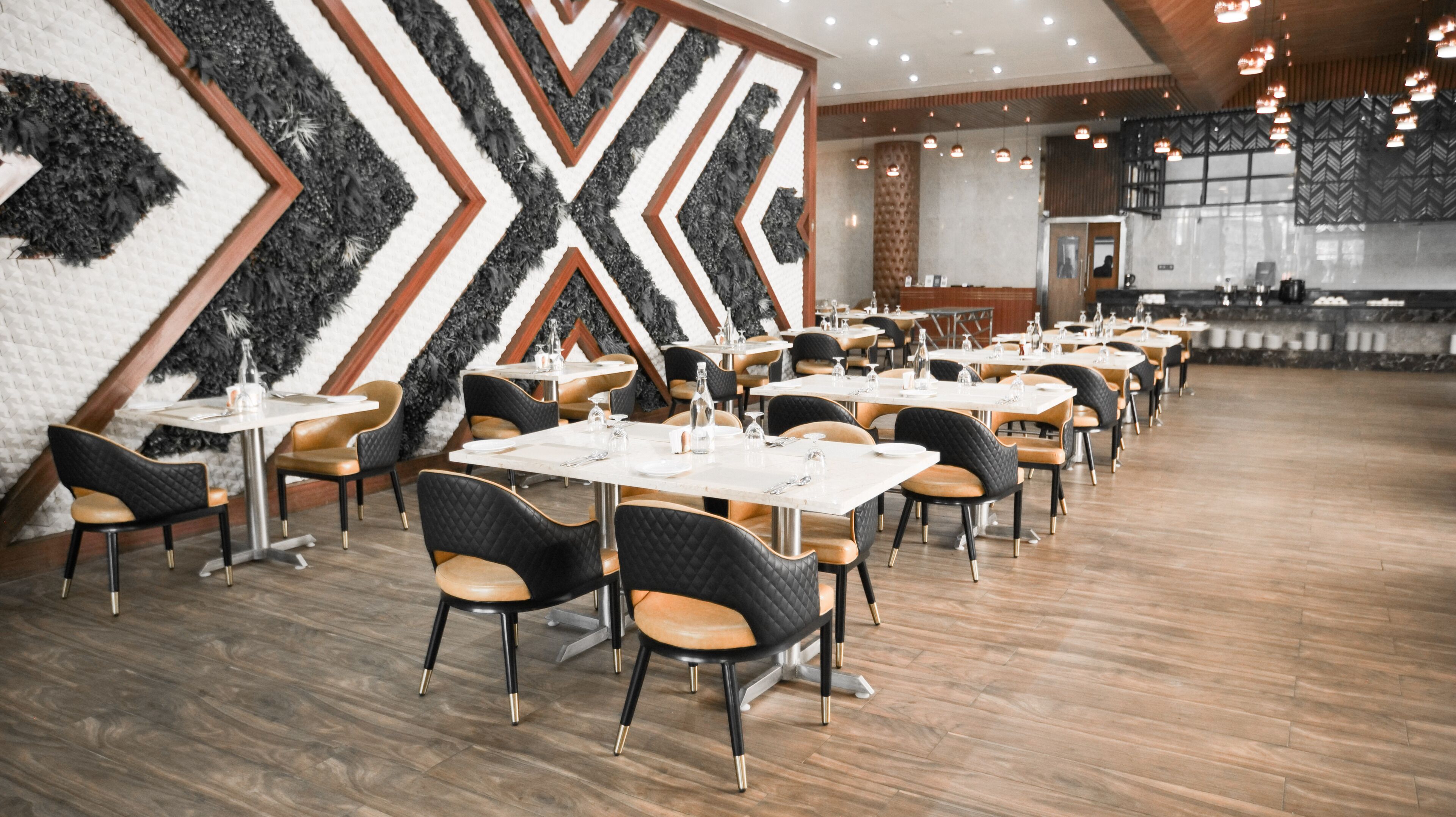 Restaurante com interior moderno, paredes com padrão preto e branco, piso de madeira e cadeiras estofadas em preto e marrom. Mesas montadas com toalhas brancas e talheres.