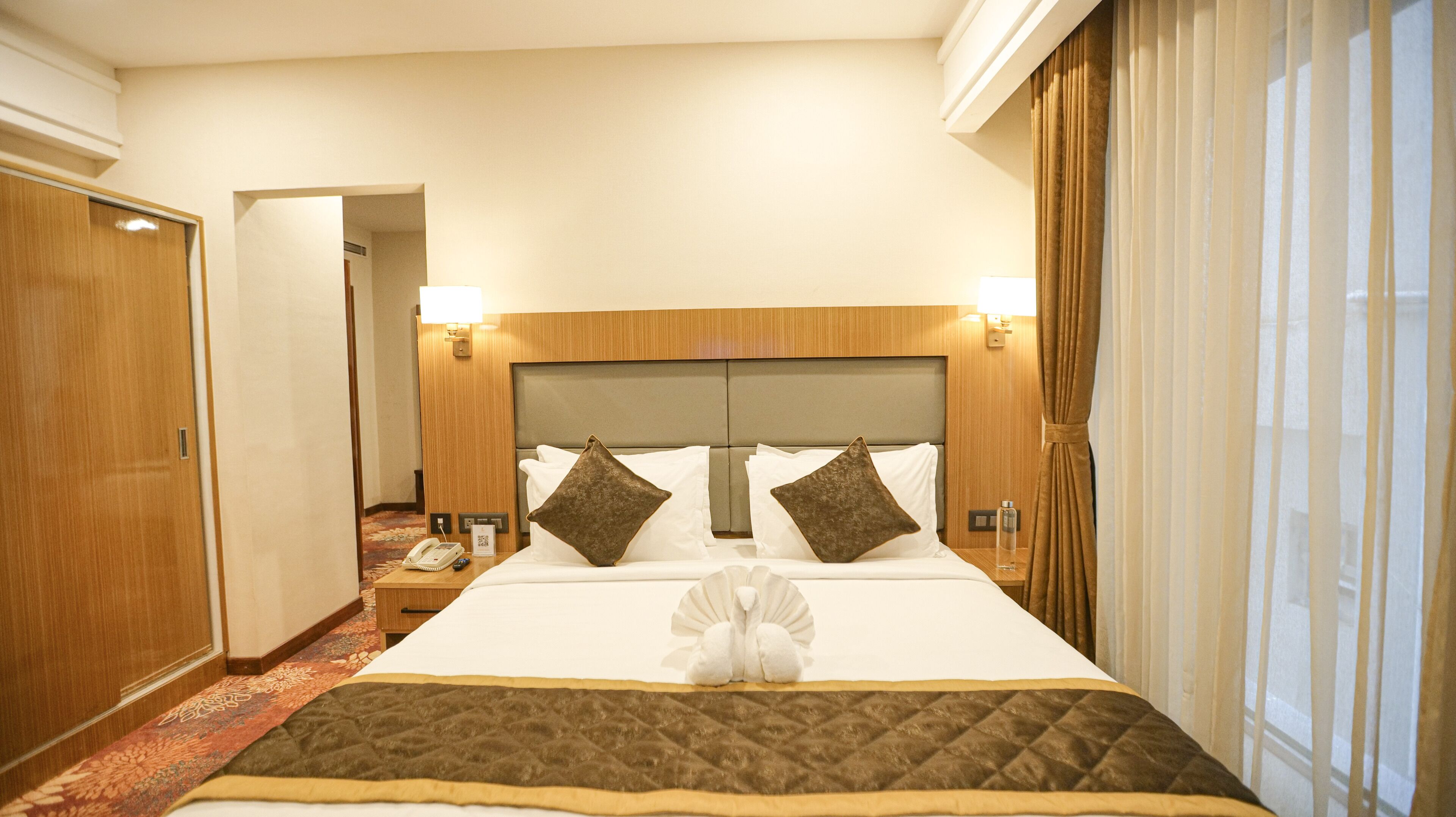 Quarto de hotel com uma cama grande, cabeceira de madeira, duas luminárias laterais, mesa de cabeceira, telefone e uma janela com cortinas.