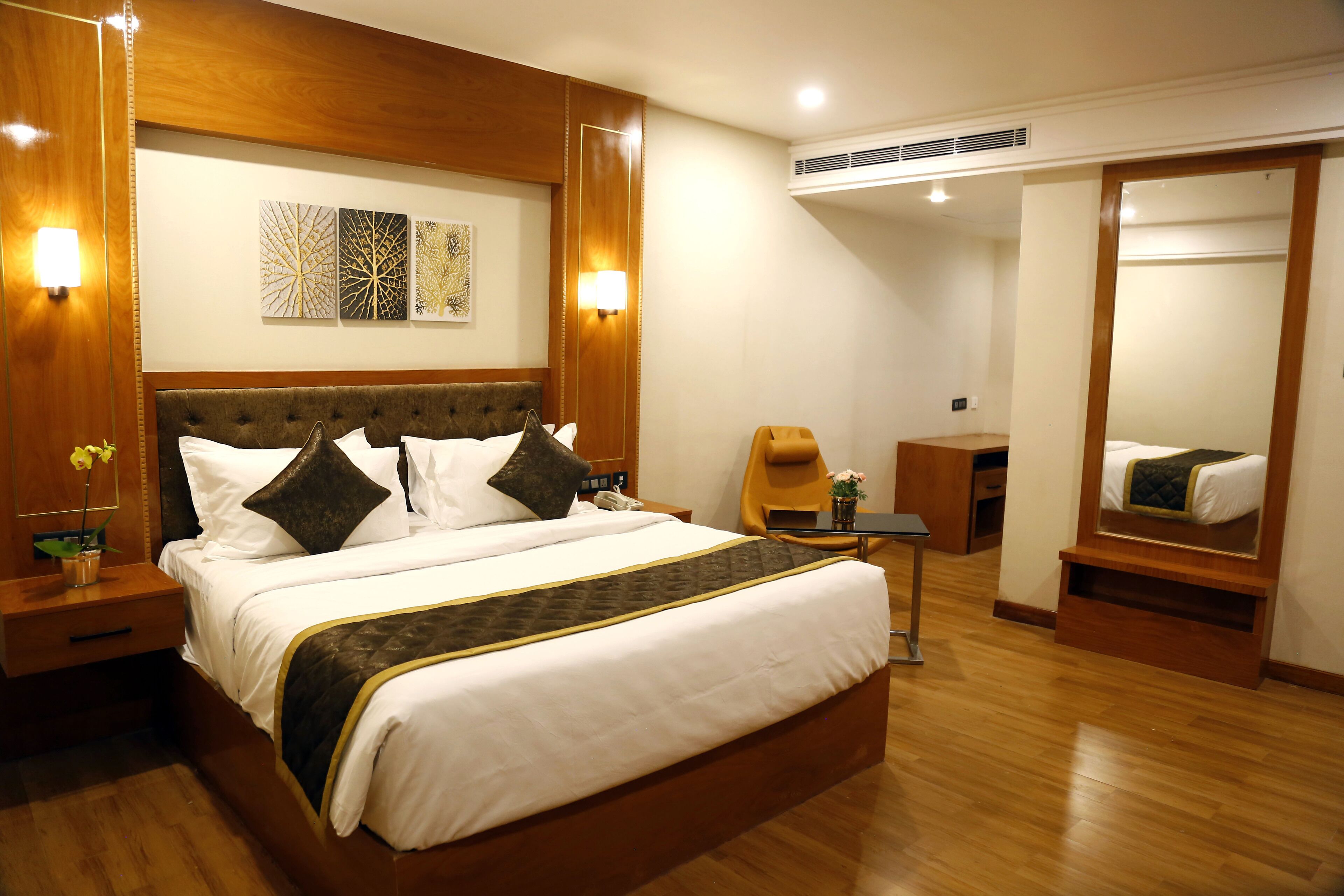 A hotel room with a large bed, wooden headboard, bedside tables, a nightstand, a mirror, a small table with a vase, and a wardrobe.