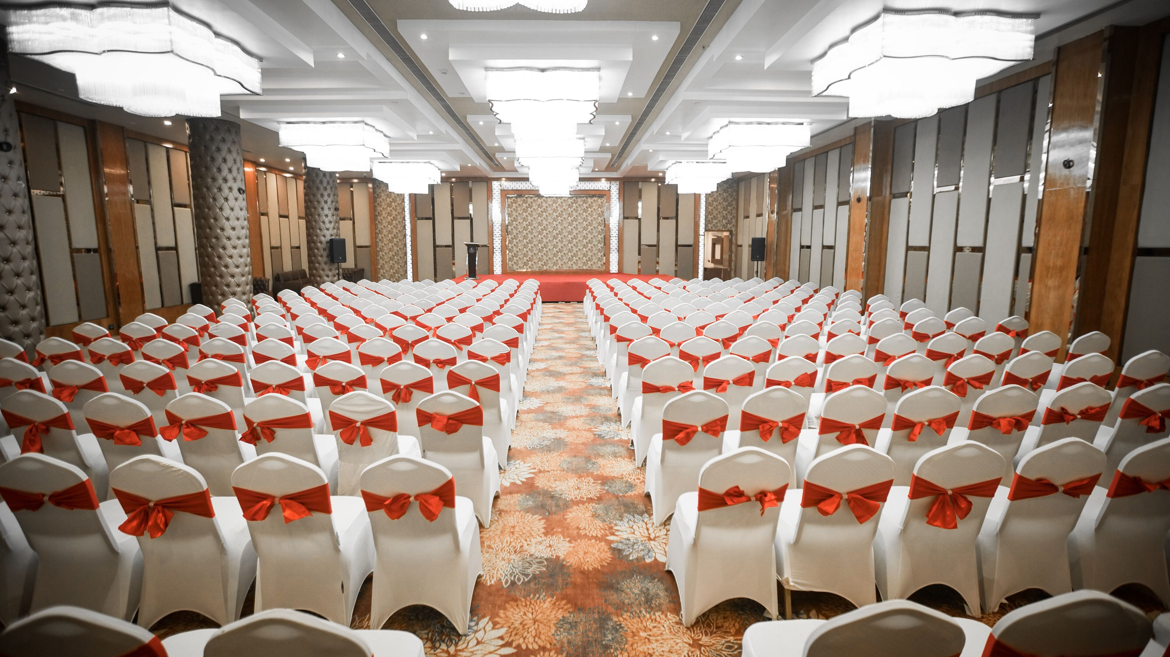 A conference room with rows of white chairs with red sashes, a patterned carpet, and a stage at the front.