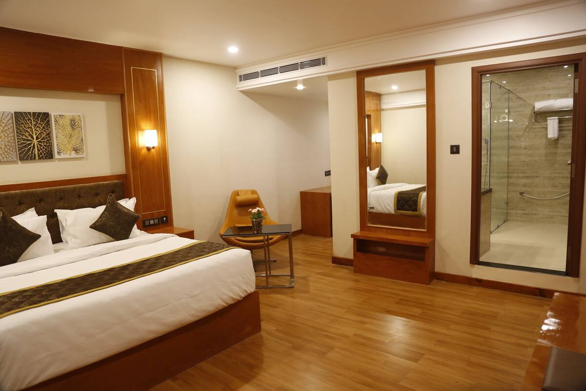 A hotel room with a large bed, a small table with a chair, a mirror, and a glass-enclosed shower area.
