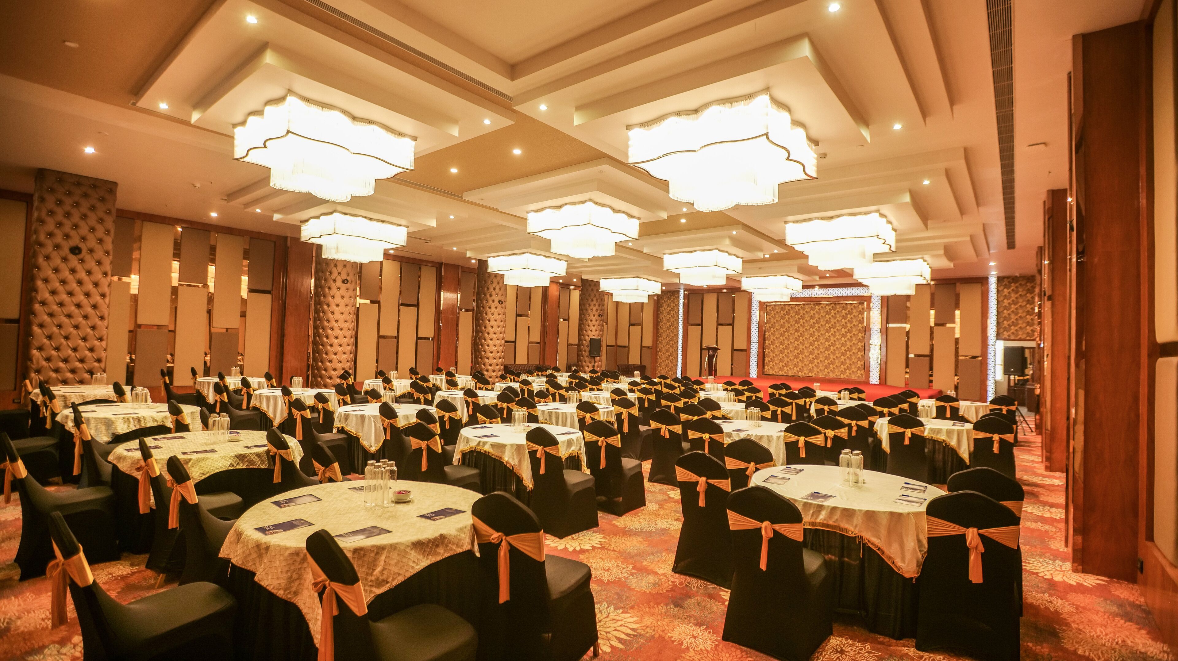 A banquet hall with multiple round tables, black chairs with orange sashes, and large chandeliers.