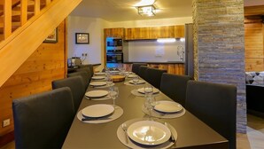 A dining area with a long table set for six, a wooden staircase, and a kitchen with wooden cabinets and a stone accent wall.