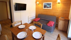Ein gemütliches Zimmer mit einer Holzwand, einem grauen Sofa mit roten Kissen, einem an der Wand befestigten Fernseher und einem Esstisch mit Tellern und Gläsern.