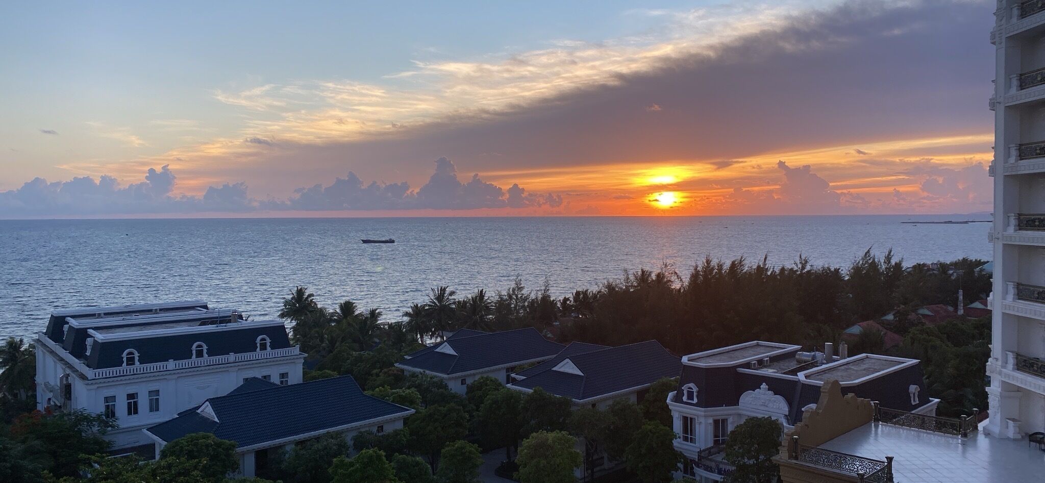 A coastal view with a sunset, buildings, and a boat on the water.