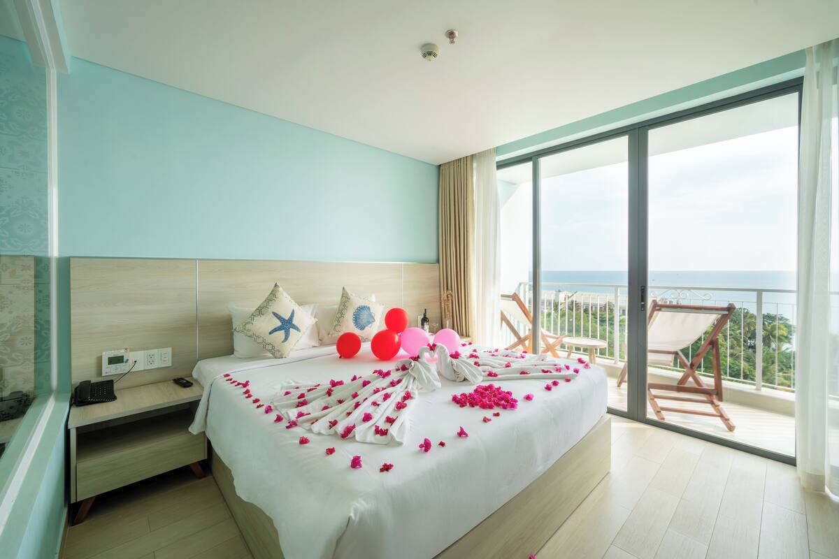 A hotel room with a large bed, a bedside table with a phone, and a view of the sea through a balcony.