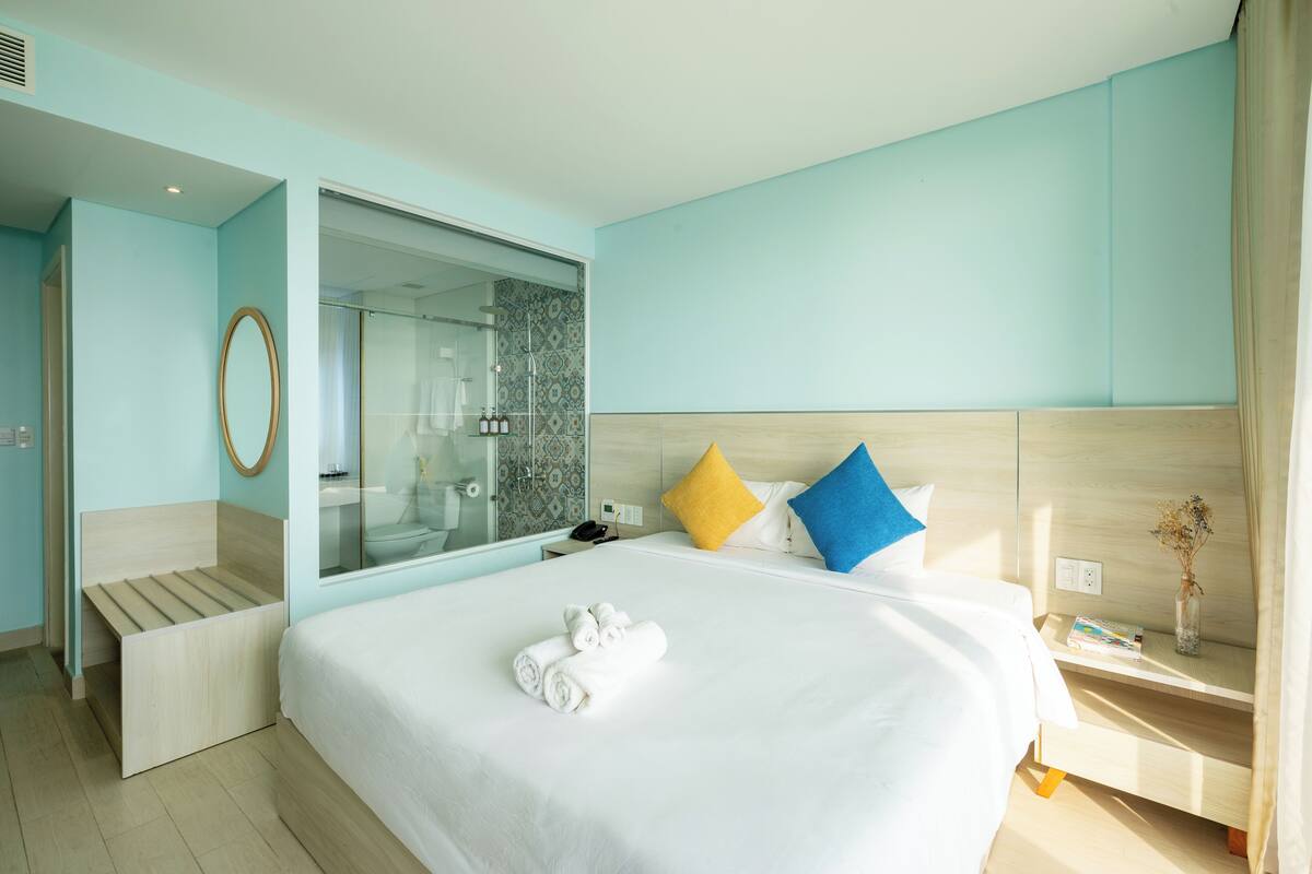 A modern hotel room with a large bed, a bedside table with a book and a vase, a mirror, and a shower area.