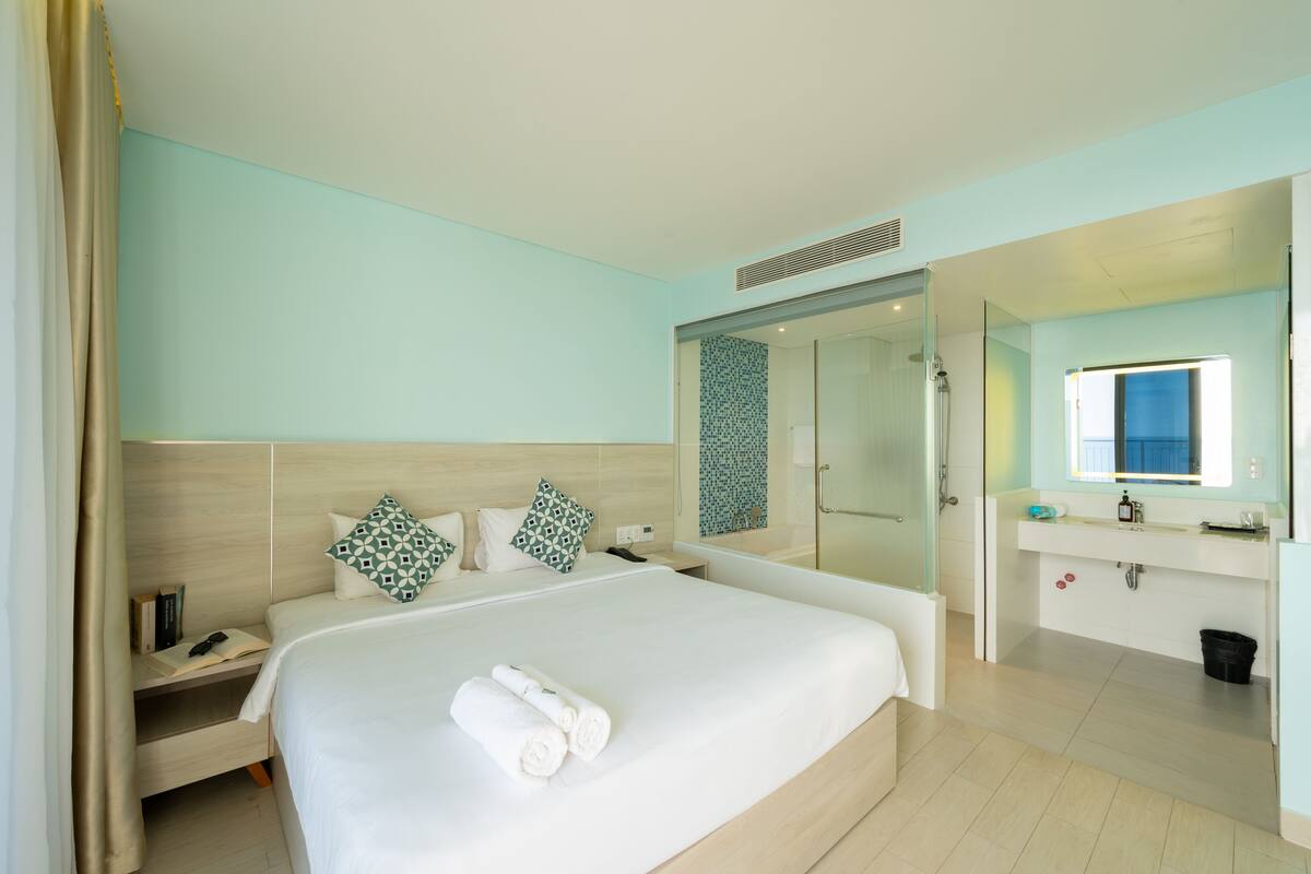 A modern hotel room with a large bed, a bedside table with a book and glasses, a bathroom with a mirror and sink, and a shower area.