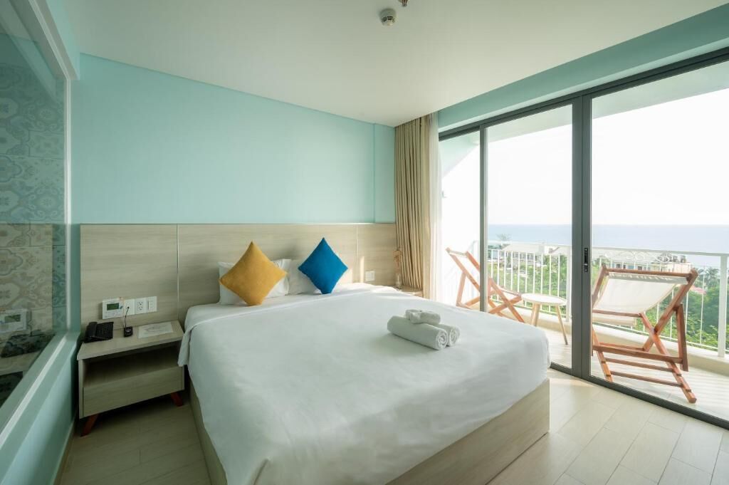 A hotel room with a large bed, a bedside table with a phone and a lamp, a window with a view of the sea, and a balcony with a chair.