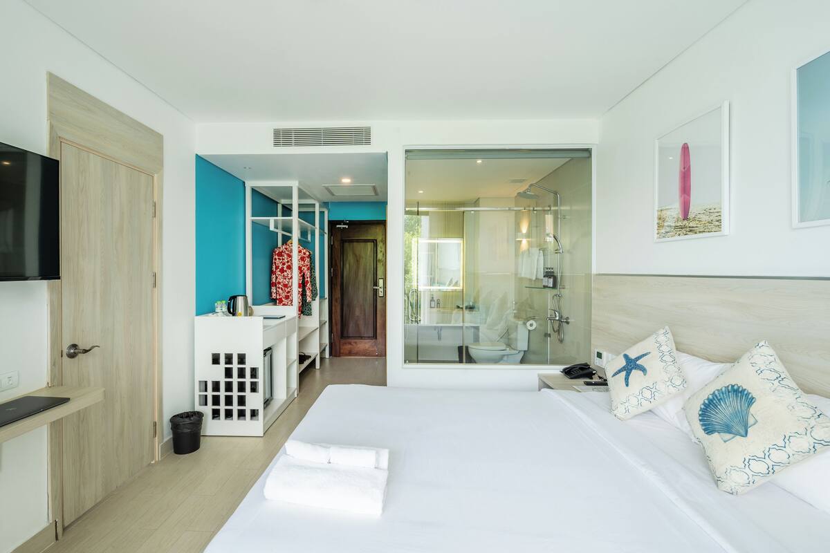 A modern hotel room with a large bed, a TV, a kitchenette, and a bathroom with a glass shower.