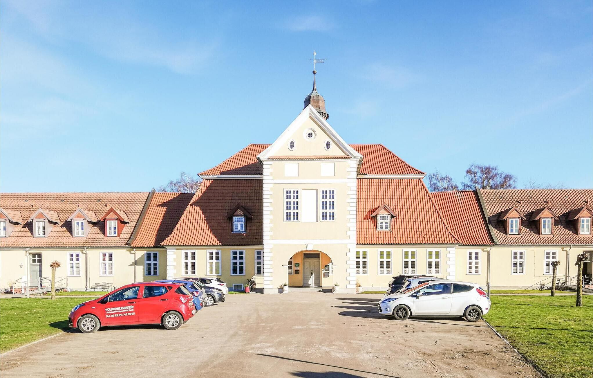 A large building with a central entrance, multiple windows, and a red-tiled roof. There are several cars parked in front.
