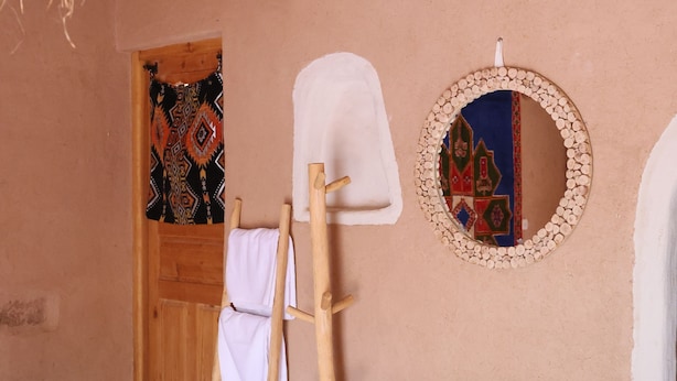Una stanza con una scala di legno, un panno bianco appeso, una porta decorata e uno specchio rotondo al muro.