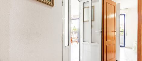 A hallway with a white wall, a framed painting, and a wooden door leading to an outdoor area with a chair and plants.