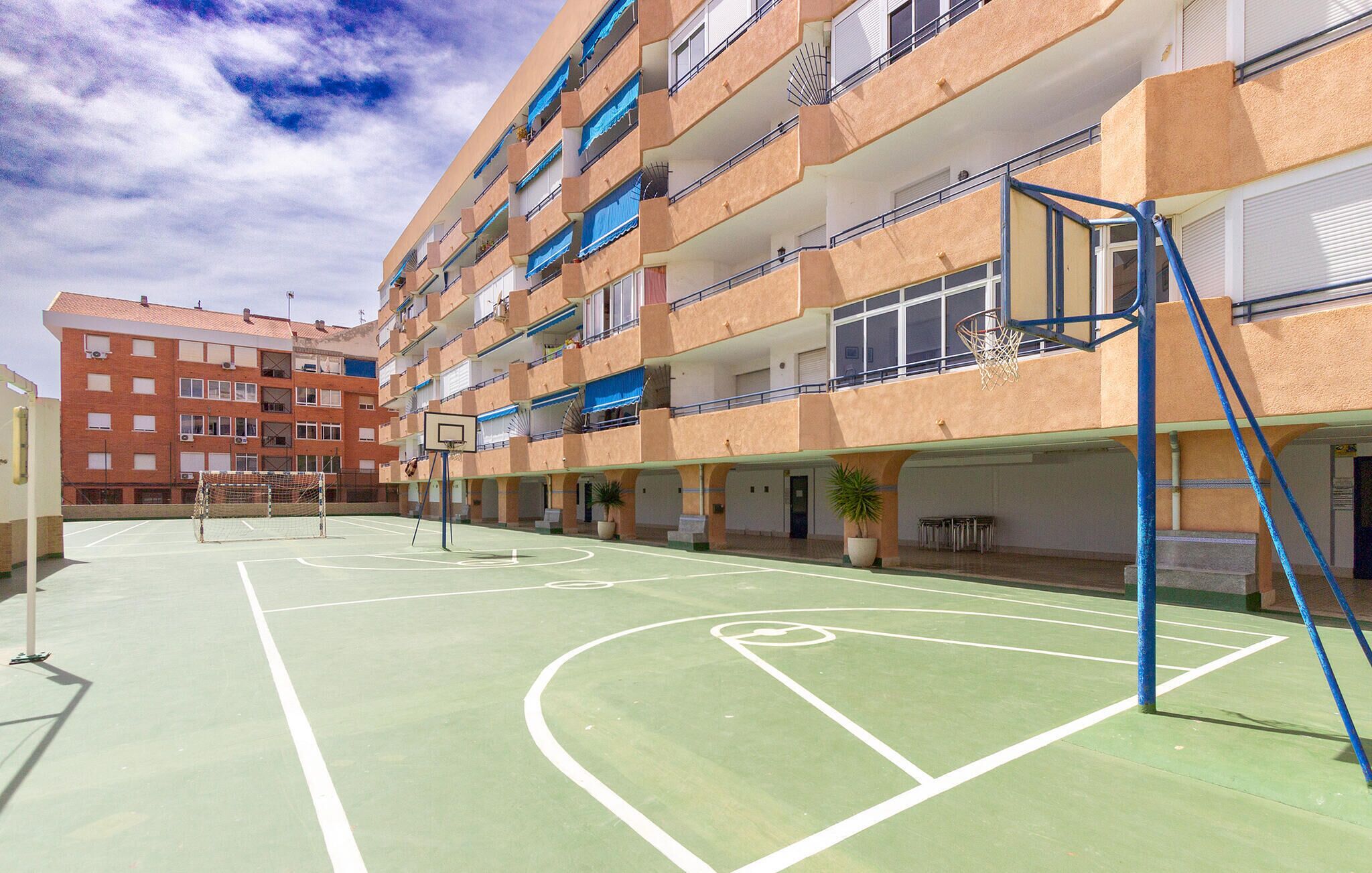 An outdoor basketball court adjacent to a multi-story residential building with balconies, a visible goal, and a clear sky.