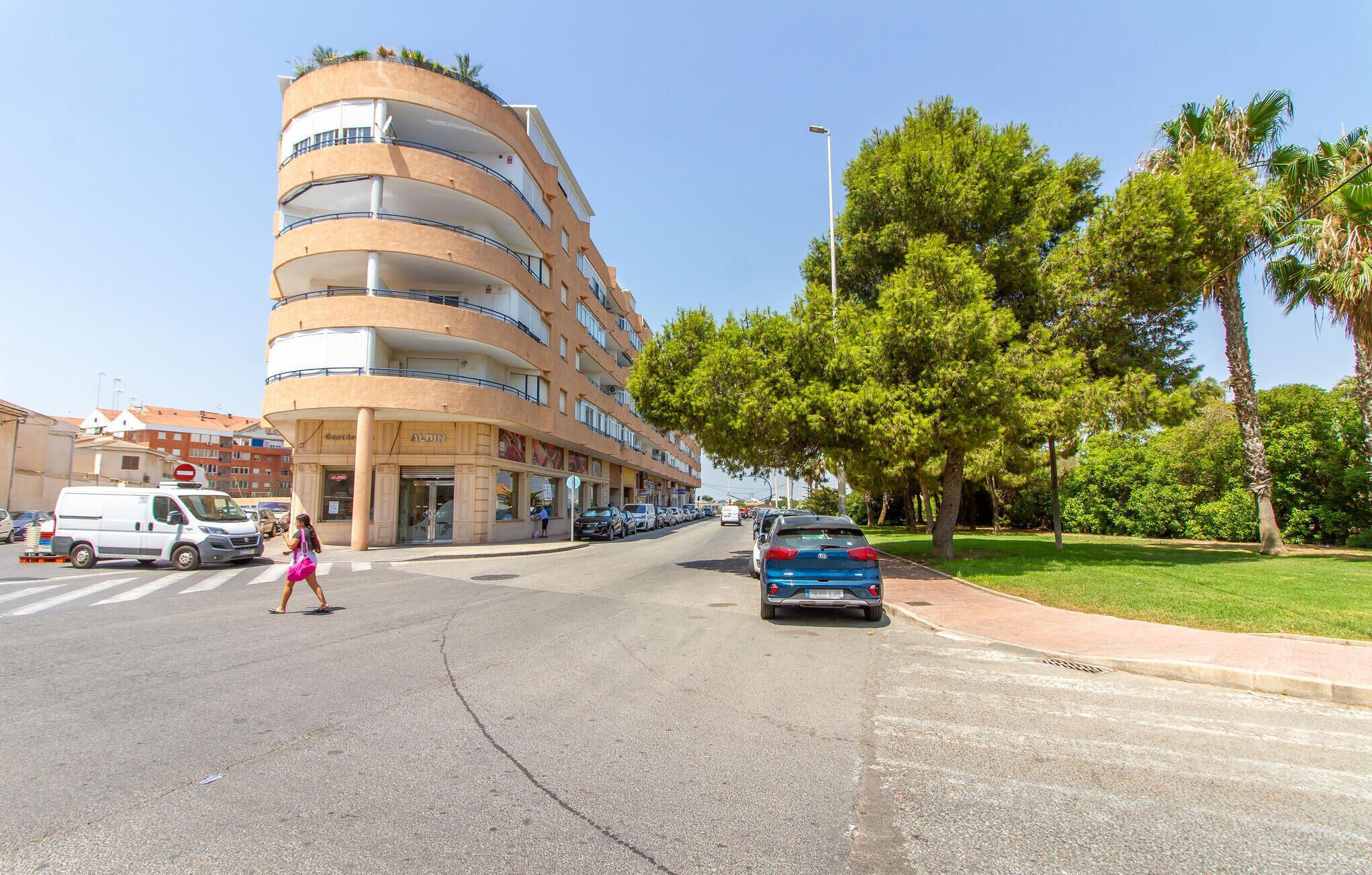 Un edificio de varios pisos con balcones, un paso de peatones y automóviles estacionados.