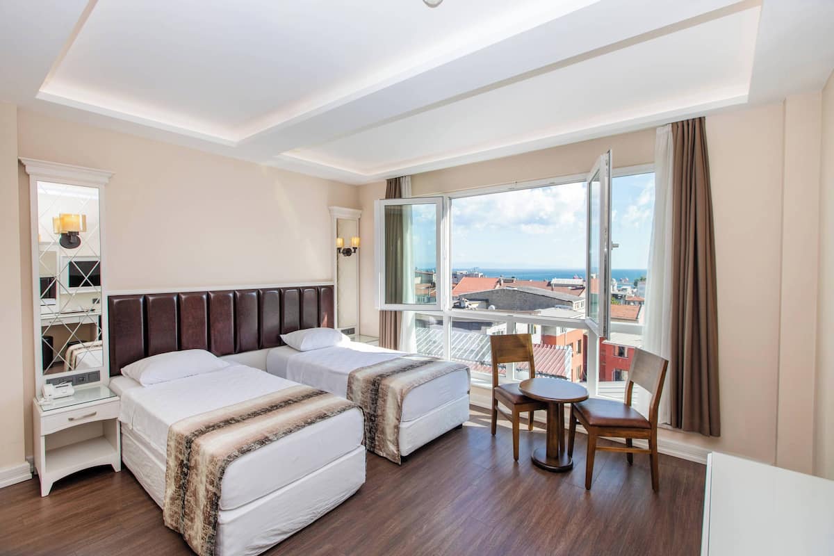 A hotel room with two beds, a small table, and a chair. There is a window with a view of the city and the sea.