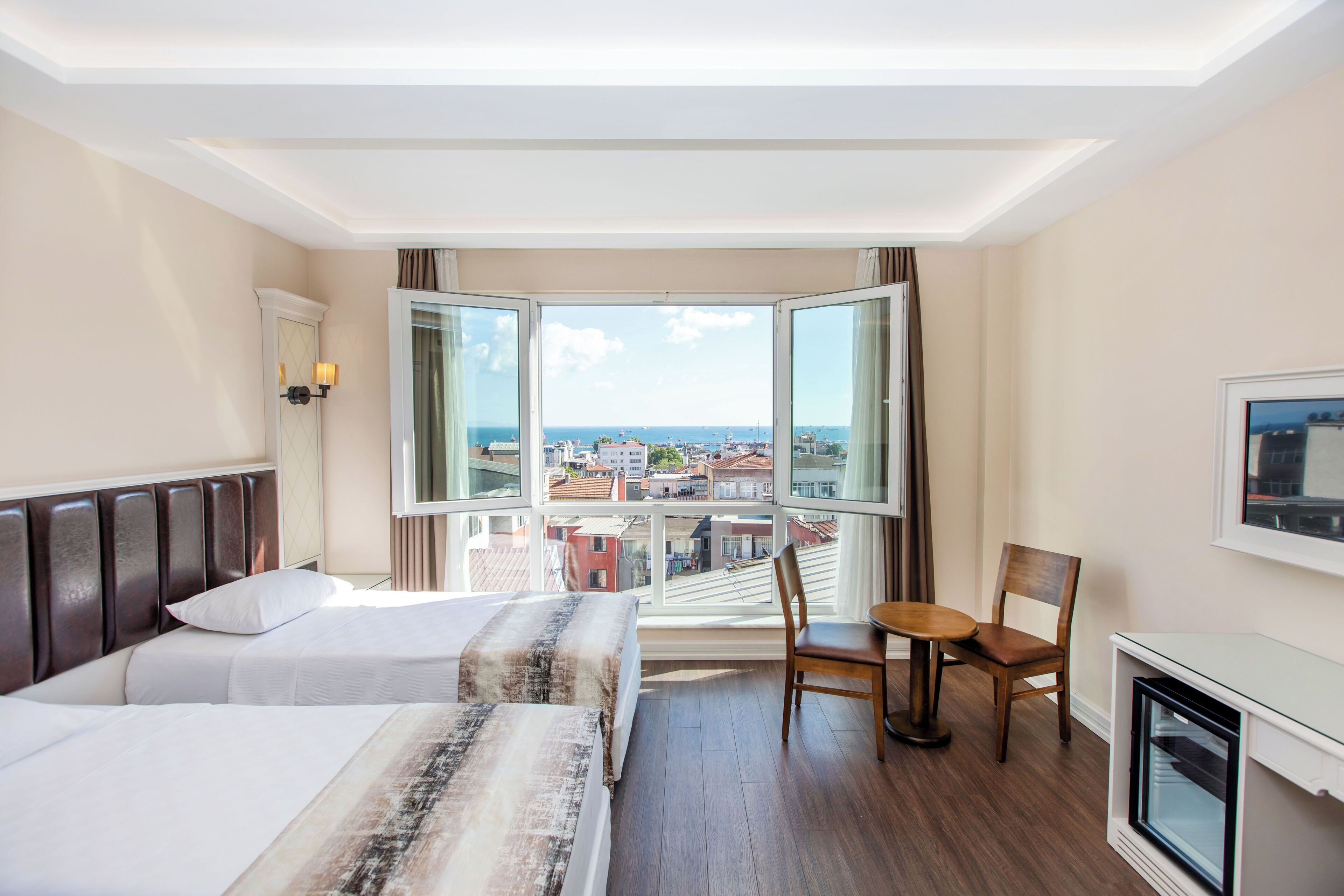 A hotel room with two beds, a small table, a chair, a TV, and a large window with a city view.