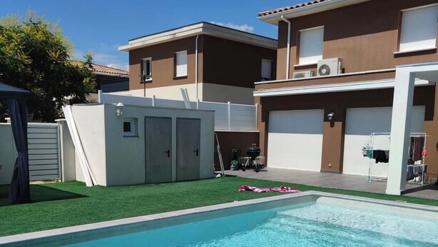 Una piscina rodeada de césped artificial, con un edificio de dos plantas al fondo.