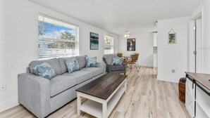 A modern living room with a grey sofa, a wooden coffee table, and a dining area with wicker chairs.