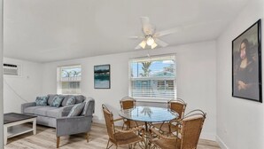 A living room with a grey sofa, a glass-top table with wicker chairs, a ceiling fan, and two windows with blinds.