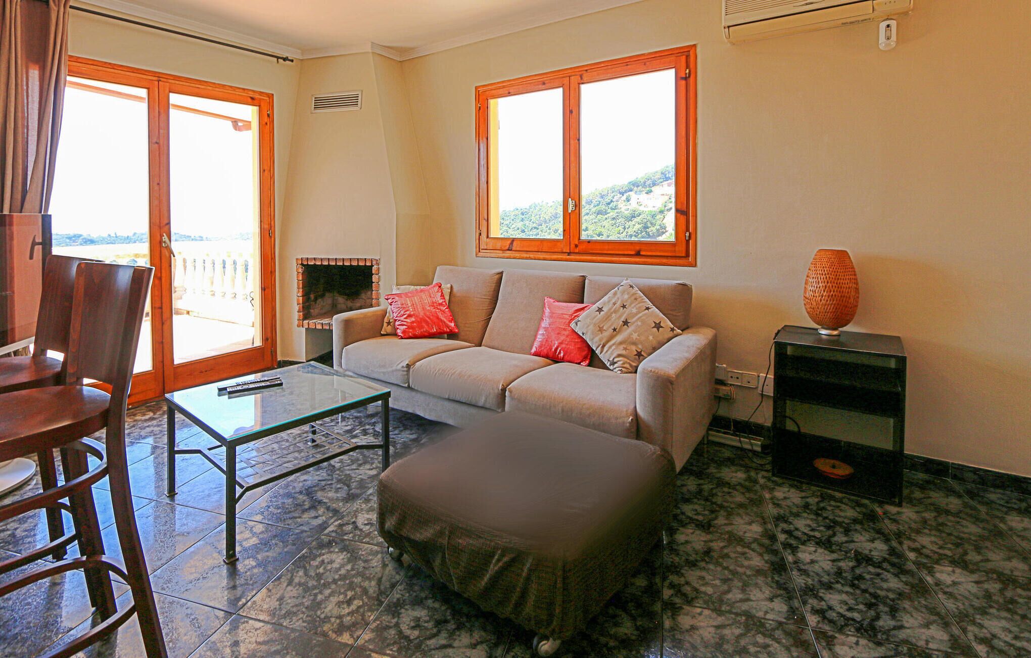 A living room with a sofa, ottoman, and a glass coffee table. There is a fireplace, a TV stand, and a window with a view of greenery.