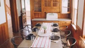 A dining area with a wooden table set for four, surrounded by chairs. There is a kitchen with wooden cabinets and a refrigerator in the background. - 5 bdrm Farmhouse. Close to the beach. (Kensington)