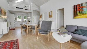 Un vaste espace de vie comprenant une table à manger, un fauteuil gris et un canapé gris. Au fond, on aperçoit une cuisine avec une rambarde blanche et, au-dessus, une mezzanine.