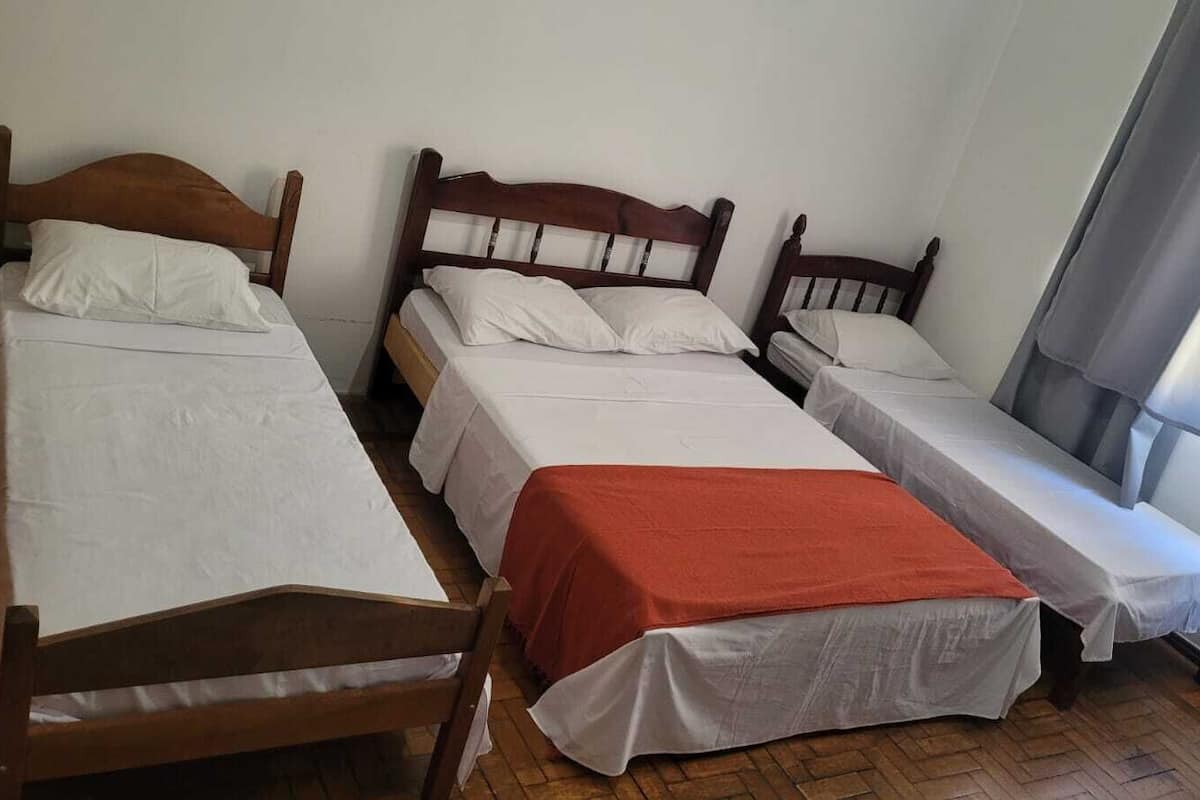 Ein Zimmer mit drei hölzernen Betten, jedes mit weißer Bettwäsche, eines davon mit einer roten Decke.