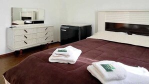 A neatly made bed with a brown bedspread, two folded white towels with green tags, a white dresser with drawers, a black appliance, and a white headboard. -  Pousada Barão Real (Sao Lourenco)