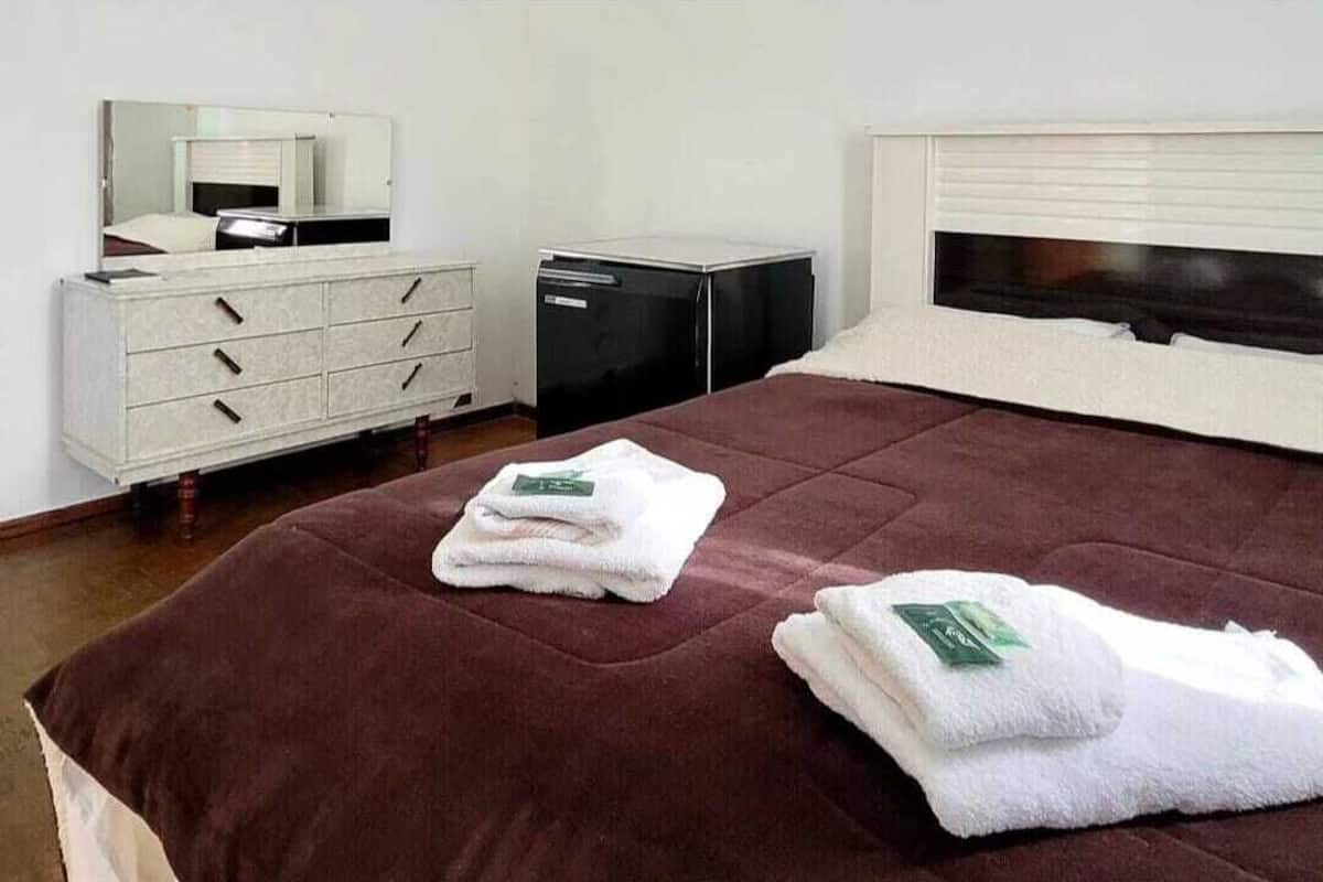 Ein ordentlich bezogenes Bett mit einer braunen Tagesdecke, zwei gefaltete weiße Handtücher mit grünen Etiketten, eine weiße Kommode mit Schubladen, ein schwarzes Gerät und ein weißer Kopfteil.