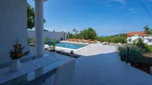 Ein moderner Poolbereich im Freien mit freiem Blick auf das Meer, umgeben von Grünpflanzen und weißen Gebäuden.