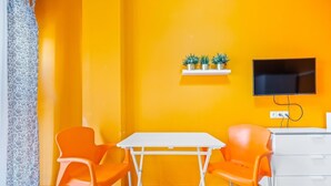 Una habitación con una pared amarilla, una mesa blanca, dos sillas naranjas, un aparador blanco y un televisor montado en la pared.