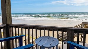 Une terrasse en bois avec une table et des chaises donnant sur une plage où l’on peut voir les vagues.