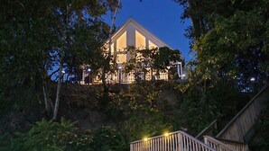 A modern building with a triangular roof, surrounded by dense foliage, and accessible by a wooden staircase.