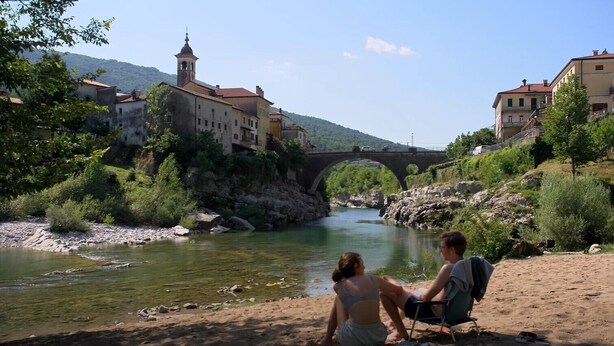 Ein Fluss mit einer Brücke, Häuser am Ufer und eine Person, die am Flussufer auf einem Stuhl sitzt.