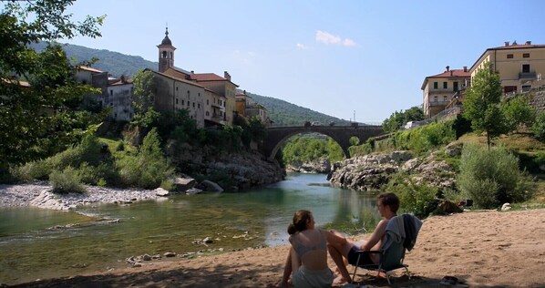 Stroll across the historic stone bridge to the tranquil riverside, surrounded by lush greenery and traditional buildings with charming balconies.
