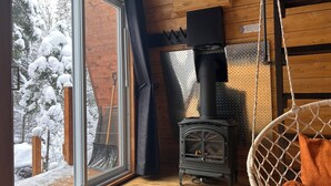Ein gemütliches Blockhaus-Interieur mit Kaminofen, Hängesessel und großem Fenster mit Blick auf eine verschneite Landschaft.