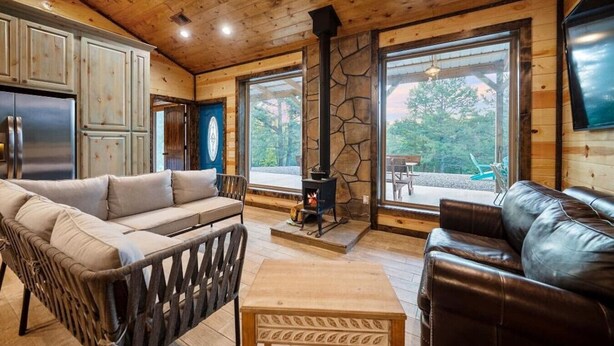 A cozy cabin interior with a stone fireplace, wooden furniture, and large windows offering a view of the outdoors. - Hidden Hills Hideaway- Broken Bow/Hochatown- 2bd (Smithville)