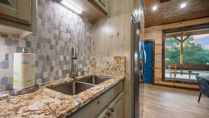 A kitchen with a granite countertop, stainless steel sink, and a refrigerator. There is a window with a view of the outdoors. - Hidden Hills Hideaway- Broken Bow/Hochatown- 2bd (Smithville)