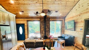 A wooden cabin interior with a dining table, chairs, a stone pillar, a ceiling fan, and a kitchen area. - Hidden Hills Hideaway- Broken Bow/Hochatown- 2bd (Smithville)